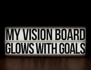 A glowing lightbox displaying the motivational message 'My Vision Board Glows With Goals' on a wooden surface.
